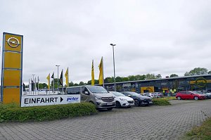 Außenansicht des Opel-Autohauses Peter in Göttingen mit moderner Glasfassade, gelbem Opel-Logo, zahlreichen geparkten Autos und einem großen Einfahrt-Schild im Vordergrund.