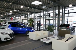 Heller, moderner Showroom eines Autohauses mit mehreren Peugeot-Fahrzeugen, unter anderem einem blauen Kleinwagen. Im Vordergrund eine Warteecke mit weißen Sesseln und Couchtisch, im Hintergrund drei Männer im Gespräch.