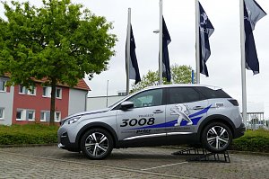 Silberner Peugeot 3008 mit Werbebeschriftung auf einer erhöhten Präsentationsrampe vor einem Autohaus. Im Hintergrund wehen mehrere Peugeot-Flaggen und es steht ein Baum sowie ein Gebäude mit roten und grauen Fassadenelementen.