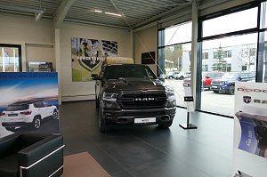 Gro&szlig;er schwarzer RAM-Pickup im Ausstellungsraum eines Autohauses mit gro&szlig;en Fenstern, daneben Werbeplakate f&uuml;r Jeep und Dodge.