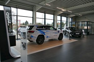 Showroom eines modernen Jeep-Autohauses mit gro&szlig;en Fensterfronten, zwei ausgestellte Jeep-Modelle, eines mit k&uuml;nstlerischer blauer Folierung, das andere in Schwarz.
