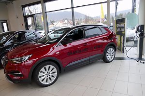 Roter Opel Grandland X Hybrid im Innenraum des Opel-Autohauses, w&auml;hrend er an einer Ladestation angeschlossen ist. Das Fahrzeug steht vor gro&szlig;en Glasfenstern, durch die man H&auml;user und H&uuml;gel im Hintergrund sieht. Auf der Fahrerseite ist die Aufschrift Grandland X Hybrid sichtbar. Daneben weitere Fahrzeuge und ein hell gefliester Boden.