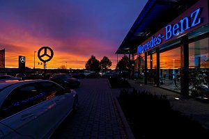 Mercedes-Benz Autohaus bei Sonnenuntergang. Der Himmel leuchtet in kr&auml;ftigen Orange- und Lilat&ouml;nen. Auf dem Parkplatz stehen mehrere Autos, der beleuchtete Mercedes-Stern und der Schriftzug �Mercedes-Benz� an der Glasfassade reflektieren das Licht.