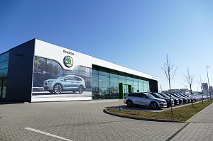 �koda-Autohaus mit großer Glasfassade und grünem Eingang, daneben viele geparkte Autos in einer Reihe, vorne ein großes Werbeplakat mit einem �koda-SUV, blauer Himmel.