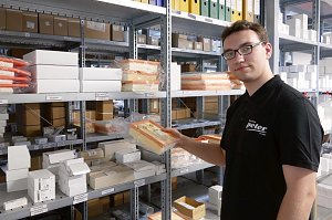 Ein junger Mann mit Brille und schwarzem Autohaus Peter-Poloshirt steht im Ersatzteillager. Er hält einen in Plastik verpackten Luftfilter in der Hand. Im Hintergrund sind zahlreiche Regale mit Autoersatzteilen, Kartons und Ordnern zu sehen.