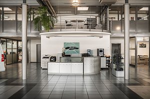 Moderner Empfangsbereich eines Autohauses mit halbkreisf&ouml;rmigem Tresen, Druckern, Kaffeemaschine und Wasserspender. An der Wand h&auml;ngt ein Bild mit Mercedes-Autos. Dar&uuml;ber eine Galerie mit Gel&auml;nder und Tisch mit St&uuml;hlen. Der Raum hat gro&szlig;e Fenster, graue Fliesen und eine hohe Decke.