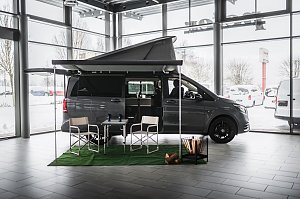 Grauer Mercedes-Benz Camper-Van mit ausgefahrener Markise und aufgeklapptem Schlafdach in einer hellen Ausstellungshalle. Vor dem Van stehen zwei Campingst&uuml;hle und ein Tisch auf gr&uuml;nem Teppich, daneben ein dekorativer Korb mit Holzscheiten. Drau&szlig;en ist Schnee zu sehen.