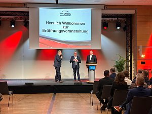 Nahaufnahme der B&uuml;hne: Drei M&auml;nner im Anzug (rechts Helmut Peter) stehen vor einer Leinwand mit der Begr&uuml;&szlig;ung zur Er&ouml;ffnungsveranstaltung. Einer spricht ins Mikrofon.