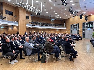 Weitwinkelaufnahme des Saals mit vielen G&auml;sten, die aufmerksam nach vorn zur B&uuml;hne schauen.