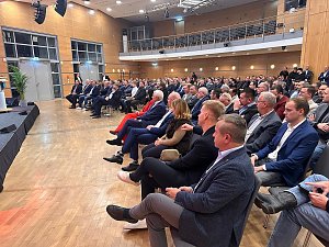 Publikum im Saal h&ouml;rt aufmerksam einer Rednerin auf der B&uuml;hne zu. Viele M&auml;nner im Anzug, einzelne Frauen im Publikum.