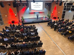 Blick von oben auf die B&uuml;hne: Prof. Dr. Mario Voigt h&auml;lt eine Rede, sein Name steht gro&szlig; auf der Leinwand.