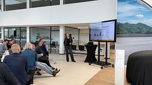 Teilnehmende verfolgen einen Vortrag im hellen �koda-Ausstellungsraum. Ein Bildschirm zeigt Diagramme, im Vordergrund Fr&uuml;hst&uuml;ckstische.