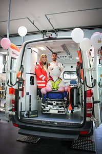 Ein ge&ouml;ffneter Rettungswagen steht im Autohaus. Zwei Mitarbeitende des Deutschen Roten Kreuzes stehen darin, vor ihnen sitzt ein gro&szlig;es Stoff-Einhorn auf einer Krankentrage. Rosa Luftballons schm&uuml;cken den Wagen.