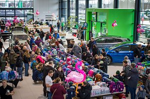 Gro&szlig;er Ausstellungsraum im �koda Autohaus, gef&uuml;llt mit vielen Menschen. In der Mitte sind lange Tische voller bunter Schulranzen. Mehrere �koda-Fahrzeuge stehen zwischen den Besuchern. Rosa Flamingo-Dekorationen h&auml;ngen im Raum.