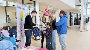 Ein Verk&auml;ufer zeigt einer Familie mit zwei Kindern einen blauen Schulranzen. Neben ihnen stehen weitere Ranzen. Im Hintergrund sind Flamingos und Schilder im Autohaus zu sehen.