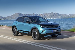 T&uuml;rkis-schwarzer Opel Mokka Electric f&auml;hrt an einer K&uuml;stenstra&szlig;e entlang, Meer und Gebirge im Hintergrund, Blick schr&auml;g von vorn links.