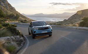 T&uuml;rkis-schwarzer Opel Mokka Electric f&auml;hrt auf einer kurvigen K&uuml;stenstra&szlig;e bergauf, Blick frontal von vorn, Meer und Berge im Hintergrund.