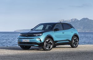 T&uuml;rkis-schwarzer Opel Mokka Electric steht am Meer, Blick schr&auml;g von vorn links, Berge im Hintergrund.