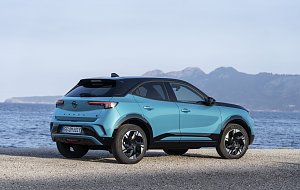 T&uuml;rkis-schwarzer Opel Mokka Electric steht am Meer, Blick schr&auml;g von hinten links, Berge im Hintergrund.