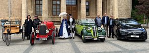 Gruppenfoto mit Fahrzeugen verschiedener Epochen: Vor einem historischen Geb&auml;ude stehen vier Autos � von einer Kutsche mit Holzaufbau &uuml;ber einen roten Oldtimer, einen gr&uuml;nen Oldtimer-Cabrio bis zu einem modernen schwarzen Mercedes. Davor posieren Personen in historischer Kleidung und moderner Garderobe.
