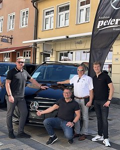 Vier M&auml;nner posieren vor einem schwarzen Mercedes Vito auf einer Kopfsteinpflasterstra&szlig;e in Heiligenstadt. Links und rechts im Hintergrund stehen weitere Fahrzeuge, im Hintergrund sind mehrst&ouml;ckige Altstadth&auml;user mit Schaufenstern zu sehen. Neben dem Auto weht eine schwarze Autohaus-Peter-Flagge. 