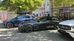 Premiumfahrzeuge in der Innenstadt: Ein blauer Mercedes-Benz SUV mit Dachbox und ein schwarzer Kombi stehen neben weiteren Fahrzeugen auf einem sonnigen Platz in einer Innenstadt mit Caf&eacute;s und Gesch&auml;ften.
