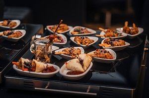Kleine wei&szlig;e Sch&auml;lchen und Gl&auml;ser mit Fingerfood, darunter Fleischspie&szlig;e, Fr&uuml;hlingsrollen und Nudeln, appetitlich angerichtet.