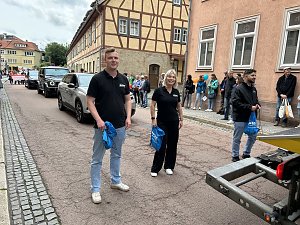 Zwei Mitarbeiter der Peter Gruppe stehen l&auml;chelnd auf der Stra&szlig;e w&auml;hrend des Festumzugs in Sondershausen. Hinter ihnen fahren Fahrzeuge der Firma, am Stra&szlig;enrand stehen Zuschauer.