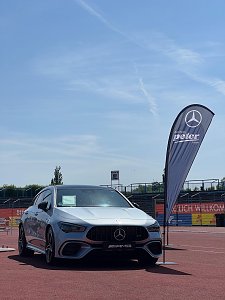 Ein silberner Mercedes-AMG steht auf der Laufbahn des Anhalt 2025 Leichtathletik-Meetings. Rechts neben dem Fahrzeug weht eine schwarze Fahne des Autohauses Peter im Wind. Im Hintergrund sind Trib&uuml;nen und Werbebanner zu sehen, w&auml;hrend der Himmel klar und sonnig ist.