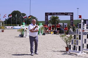Helmut Peter spricht mit einem Mikrofon auf dem Springplatz beim Reitturnier in Immenrode. Hinter ihm stehen Helfer in roten Shirts vor dem Begr&uuml;&szlig;ungsbogen und wehenden Fahnen.