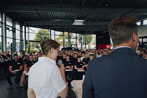 Blick von hinten auf Lilly Hecker und Gesch&auml;ftsf&uuml;hrer Andreas Peter, w&auml;hrend sie vor den neuen Auszubildenden sprechen. Viele junge Menschen sitzen in Reihen und h&ouml;ren zu.