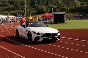 Nahaufnahme des weißen Mercedes-Cabrios auf der Laufbahn. Drei Sportstars sitzen auf der Rückbank, jubeln und winken dem Publikum zu.