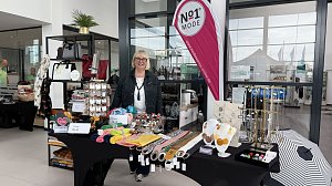 Stand von No1 Mode im Autohaus Autowelt Peter. Eine Frau pr&auml;sentiert Taschen, T&uuml;cher, Schmuck und Accessoires, die auf Tischen und Regalen dekoriert sind. Daneben steht eine pinkfarbene Werbefahne mit dem Logo des Modegesch&auml;fts.