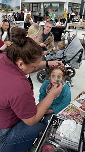 Kinderschminken im Autohaus Autowelt Peter: Ein M&auml;dchen mit Katzenohren-Stirnband bekommt ein buntes Regenbogen-Gesicht gemalt. Im Hintergrund sitzen weitere Familien mit Kindern.