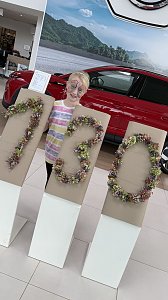 Ein Kind mit bunt bemaltem Gesicht steht im Autohaus Autowelt Peter hinter gro&szlig;en Ziffern aus Blumen, die �130� formen � passend zum Jubil&auml;um 130 Jahre �koda. Daneben ist ein rotes Auto ausgestellt.