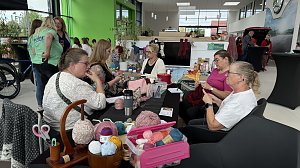 Frauen sitzen an einem Tisch im Autohaus Autowelt Peter und stricken gemeinsam. Auf dem Tisch liegen bunte Wollkn&auml;uel, Strickutensilien und Handarbeiten. Im Hintergrund sind weitere G&auml;ste und Ausstellungsst&auml;nde zu sehen.