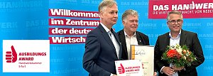 Drei M&auml;nner stehen vor einer blauen Wand mit dem Schriftzug Das Handwerk � Die Wirtschaftsmacht von nebenan. Einer h&auml;lt eine Urkunde, ein anderer eine Blumenstrau&szlig;, der dritte das Schild Ausbildungsaward 2025 Handwerkskammer Erfurt. Das Bild zeigt die &Uuml;bergabe des Ausbildungsawards an die Autohaus Peter Gruppe.
