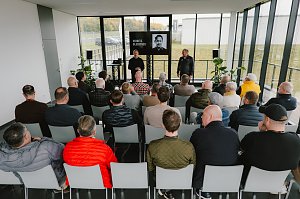 Eine Gruppe von Menschen sitzt in einem modernen, hellen Raum während einer Präsentation. Im Vordergrund sieht man mehrere Männer, die auf Stühlen sitzen und auf eine Bühne blicken. Auf der Bühne stehen zwei Personen: Ein Mann trägt einen schwarzen Pullover und spricht, während der andere in einem schwarzen Outfit steht. Auf einem Bildschirm im Hintergrund wird ein Porträt von Moritz Bleibtreu gezeigt. Der Raum hat große Fenster, durch die man die Natur draußen sieht. Pflanzen stehen in den Ecken des Raumes.
