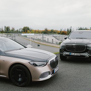 Zwei Luxusautos von Mercedes-Benz stehen nebeneinander auf einer Rennstrecke. Auf der linken Seite ist ein silberner Mercedes-Maybach S-Class zu sehen, der eine glänzende schwarze Motorhaube und schwarze Räder hat. Rechts daneben steht ein schwarzer Mercedes-Maybach GLS, ein großer SUV mit auffälligem Kühlergrill und silbernen Akzenten. Im Hintergrund erstreckt sich die Rennstrecke, und Bäume mit herbstlichen Farben sind am Horizont sichtbar.