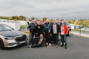 Eine Gruppe von Männern steht lachend zwischen zwei Mercedes-Maybach Fahrzeugen auf dem Gelände der Mercedes-Benz Driving Experience am Bilster Berg. Einige tragen bunte Jacken, und einer sitzt entspannt auf dem Boden. Im Hintergrund sind Gebäude der Rennstrecke und herbstlich gefärbte Bäume zu sehen.