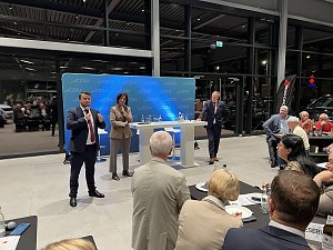 Drei Personen stehen vor einem CDU-Hintergrund auf einer B&uuml;hne im Autohaus. Eine Frau und zwei M&auml;nner sprechen in Mikrofone, w&auml;hrend das Publikum aufmerksam zuh&ouml;rt. Auf den Tischen stehen Wasserflaschen und Gl&auml;ser.