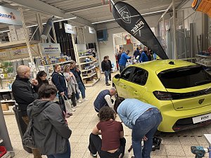 Gelber Peugeot im Baumarkt; viele interessierte Baumarktbesucher stehen rund herum und sehen beim Räderwechsel zu.
