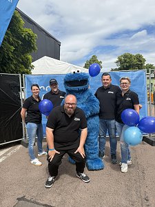 Man sieht 5 Mitarbeiter des Autohaus Peter zusammen mit dem Kr&uuml;melmonster aus der Sesamstra&szlig;e mit blauen Luftballons vor einer blauen Bauzaunplane.