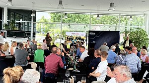 Autohaus mit vielen Personen an Tischen sitzend; vorne spricht ein Mann ins Mikrofon.