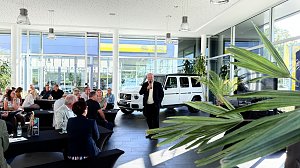 Autohaus mit vielen Personen an Tischen sitzend; vorne spricht ein Mann ins Mikrofon, dahinter ein weißes Mercedes-Fahrzeug.