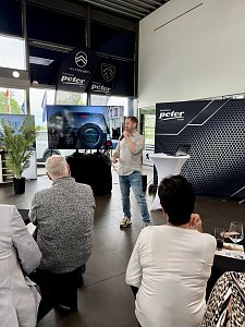 Eine Person steht vorn und spricht ins Mikrofon, im Hintergrund ein Bildschirm.