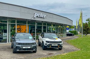 Außenansicht des Autohauses Peter in Halle Neustadt/ Göttinger Bogen. Im Vordergrund steht ein grauer und ein weißer Jeep. Rechts stehen Fahnenmasten auf einer Grünfläche. Im Hintergrund ist das Autohaus mit einer großen Glasfront.