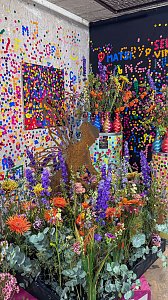 Eine farbenfrohe Dekoration mit üppigen, bunten Blumenarrangements im Vordergrund vor Wänden, die mit bunten Punkten und abstrakten Mustern im Pop-Art-Stil beklebt sind.