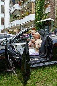 Zwei Frauen, die lächelnd in einem schwarzen Mercedes-Cabriolet mit geöffnetem Verdeck sitzen und mit Getränken anstoßen.