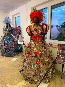 Zwei kunstvoll gestaltete Schaufensterpuppen, die in einem Innenraum aufwendige, voluminöse Kleider aus ungewöhnlichen Materialien präsentieren, wobei das vordere Kleid von roten Herzen und floralen Elementen geprägt ist.